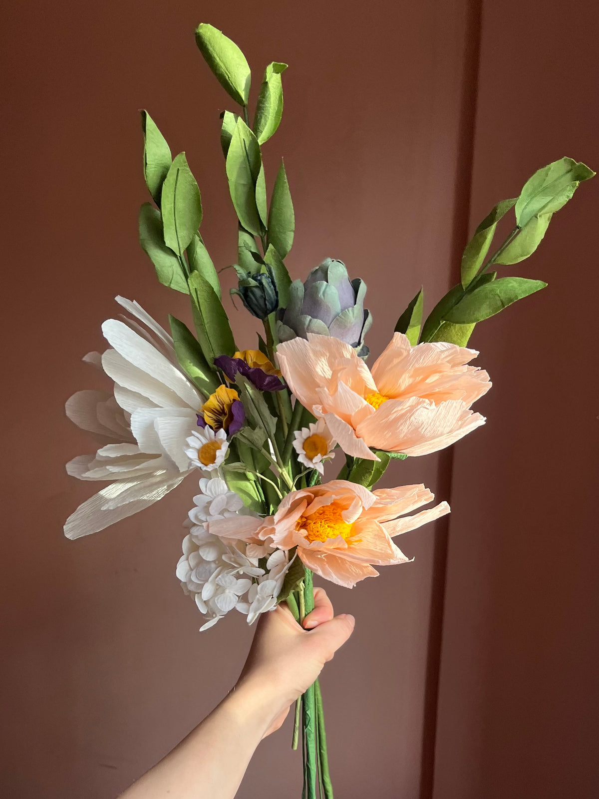 Hånd holder evighedsbuket af papirblomster med ferskenfarvede blomster og artiskok
