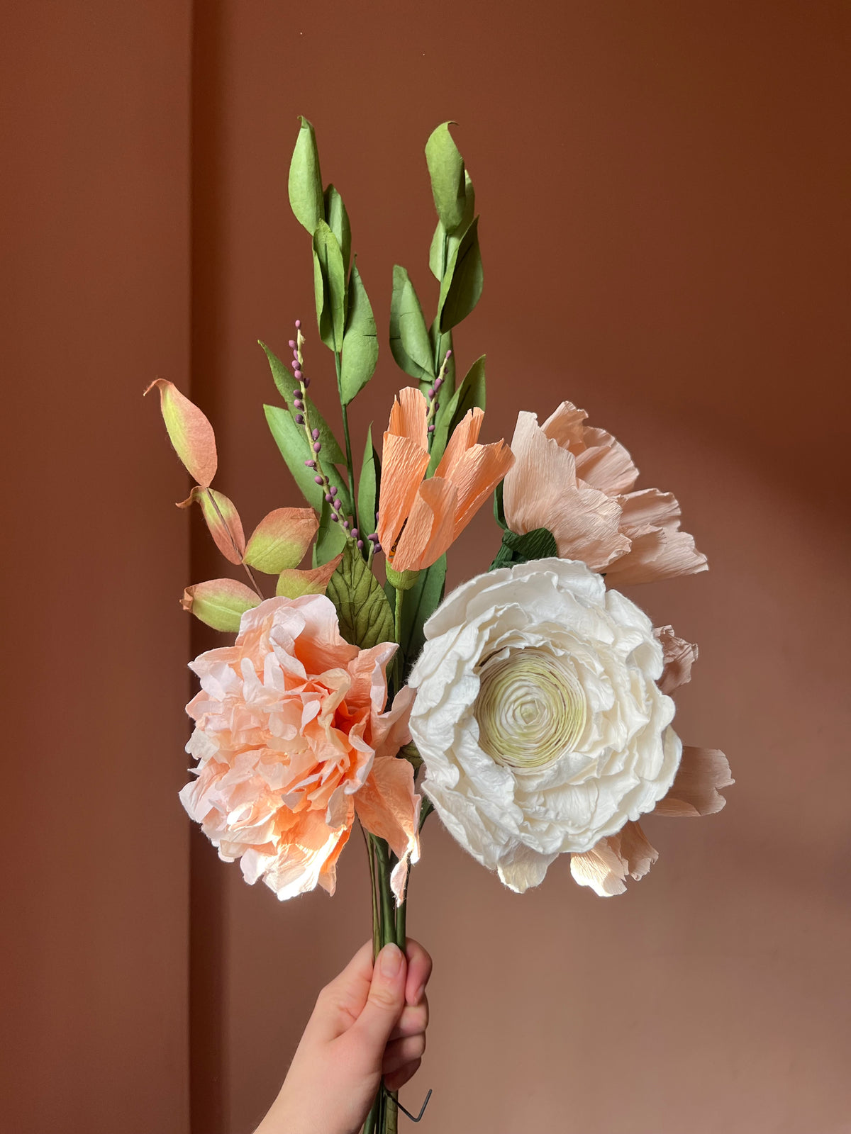 Hånd holder evighedsbuket af papirblomster med creme ranunkel og ferskenfarvede valmuer