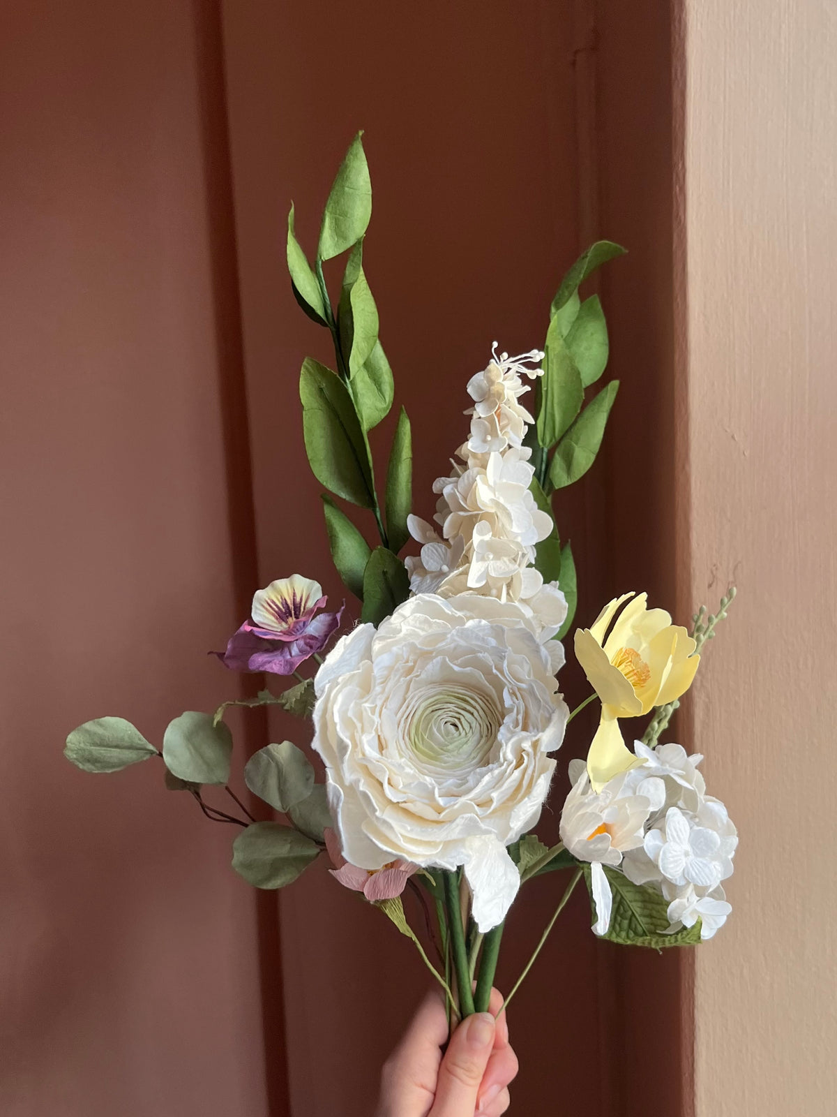 Hånd holder hvid evighedsbuket af papirblomster med lysegul blomst og stedmoderblomst