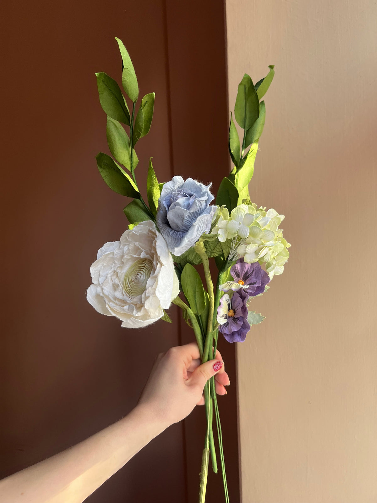 Evighedsbuket af papir med hvid ranunkel, blå rose og lilla stedmoderblomster mod lyserød væg