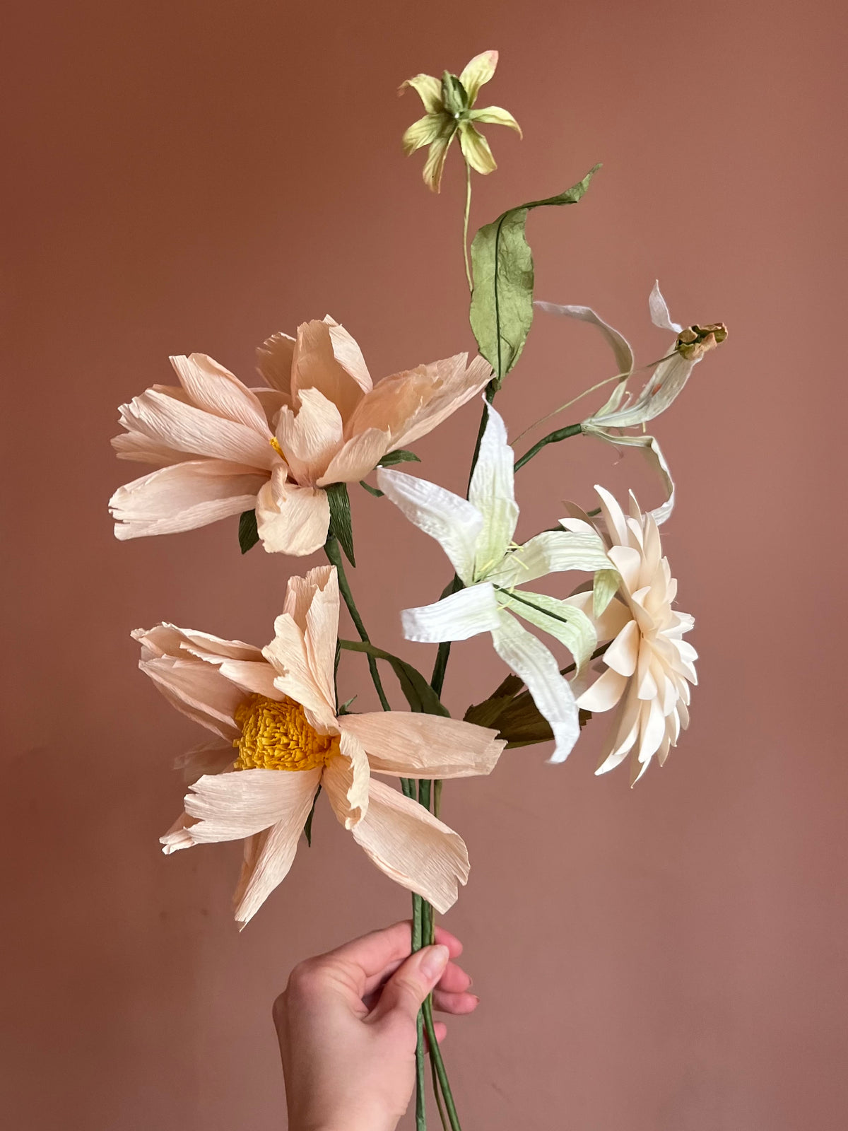 Hånd holder evighedsbuket af papirblomster med ferskenfarvede blomster, creme dahlia og lilje